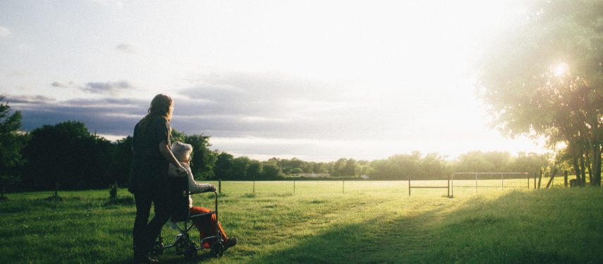 A young woman pushes an older person in a wheelchair in a field where the sun is setting