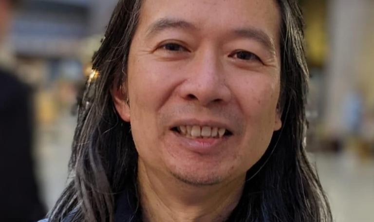 head and shoulders shot of Andrew Chiu, he is an asian man and has long hair and is wearing a blue top, he's smiling at the camera