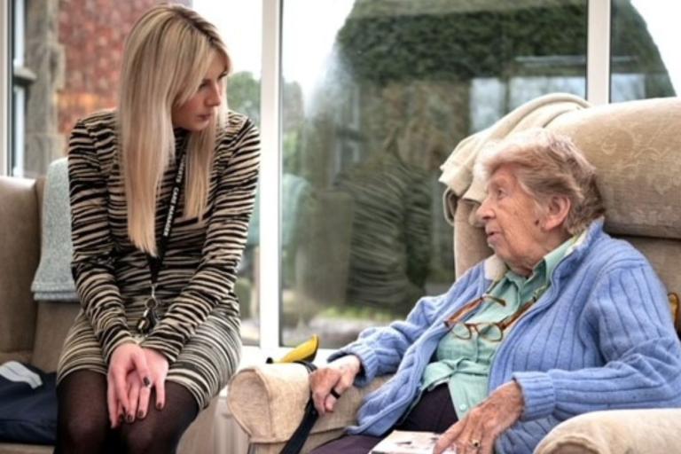 Young woman sat on the edge of a chair talking to an older woman sat in a chair at a care home