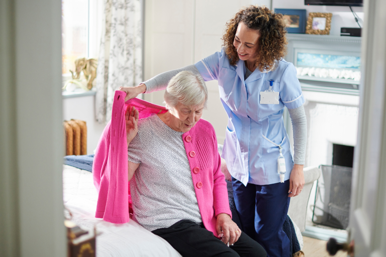 An older woman sat on a bed being helped by a carer to put a cardigan on 